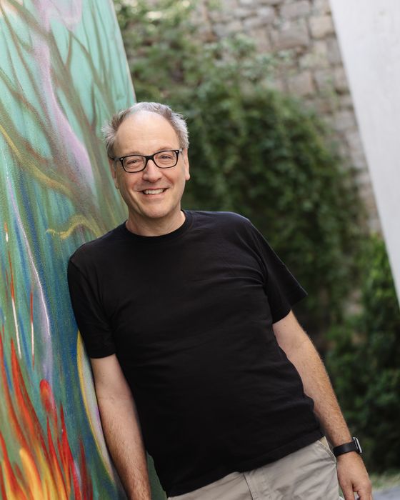 Stephan Schug se usmívá do objektivu. Opírá se o barevně pomalovanou zeď, má brýle a černé tričko.